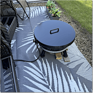 Round black portable charcoal grill with lid sits on a palm-patterned outdoor rug near wicker chairs in a patio setting.