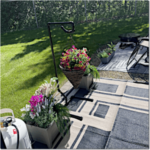 Backyard patio with a hanging flower basket and planter boxes on a patterned outdoor rug; a propane tank is nearby and a seating area is in the background.