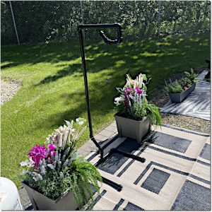 Black metal plant stand on a patterned outdoor rug, flanked by two rectangular planters with white and purple flowers; a green lawn and chain-link fence in the background.