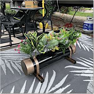 Black cylindrical planter with copper end caps and leather straps holding a mix of green and flowering plants on a patterned outdoor rug.