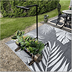 Plant cart with a grow-light over potted greenery on a gray palm-leaf patterned outdoor rug beside a gravel path and patio furniture.